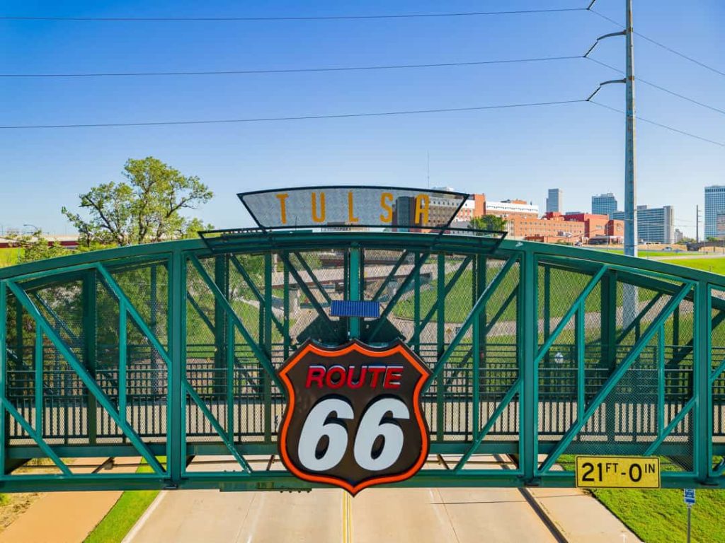 Groene stalen brug met een verlicht bord met de tekst tulsa route 66, met de skyline van Tulsa op de achtergrond