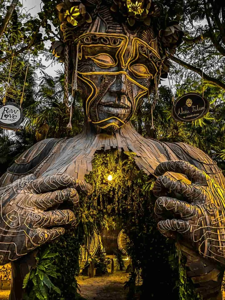 Een groot houten beeld met een Maya gezicht en uitgestoken handen vormt de poort naar een strandclub in Tulum