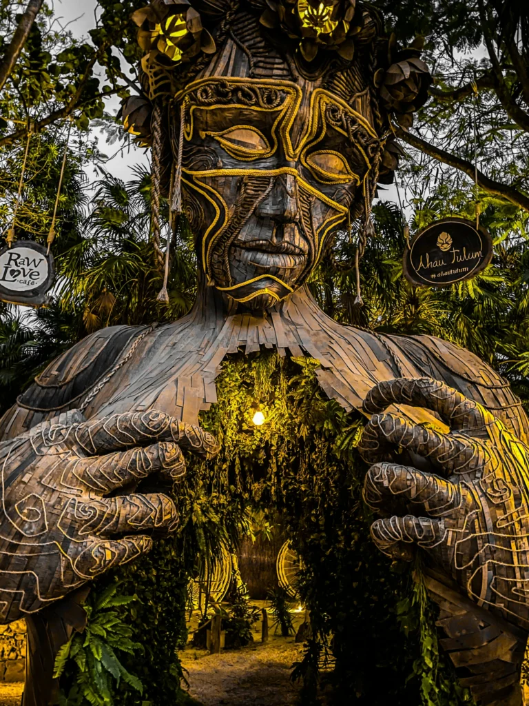Een groot houten beeld met een Maya gezicht en uitgestoken handen vormt de poort naar een strandclub in Tulum