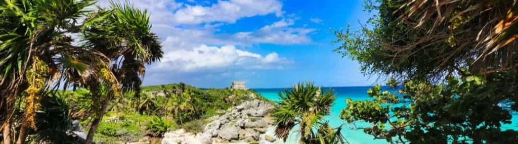Doorkijkje tussen palmbomen naar de ruïnes van Tulum op een klif boven een turquoise zee