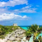 Doorkijkje tussen palmbomen naar de ruïnes van Tulum op een klif boven een turquoise zee