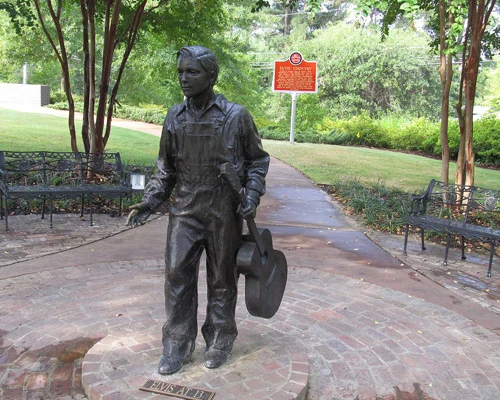 Bronzen standbeeld van een tienerjongen in overall met een gitaarkoffer in een park in Tupelo in Mississippi, met een historisch informatiebord op de achtergrond