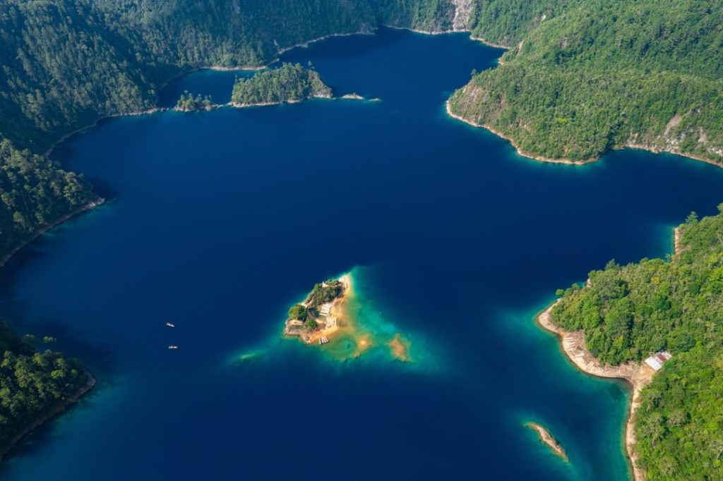 Luchtfoto van een dieppaars blauw meer met een zandkleurig eilandje omringd door dicht groen bos
