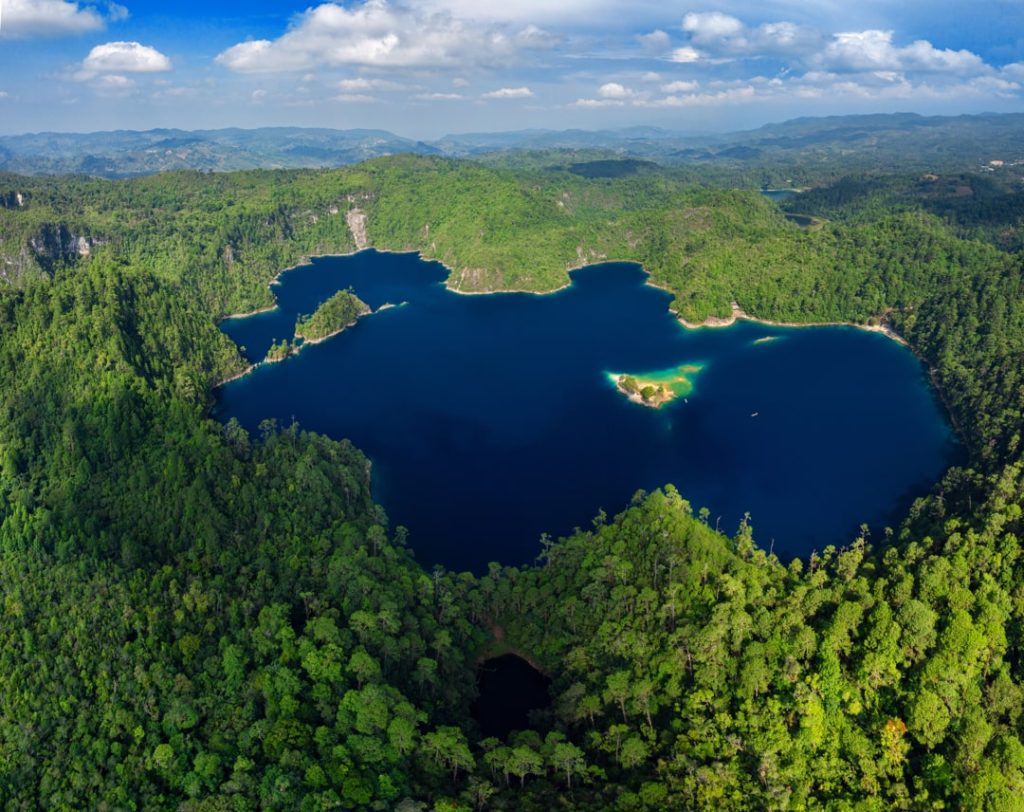 Luchtfoto van een diepblauw meer met een klein begroeid eilandje omringd door dicht tropisch bos