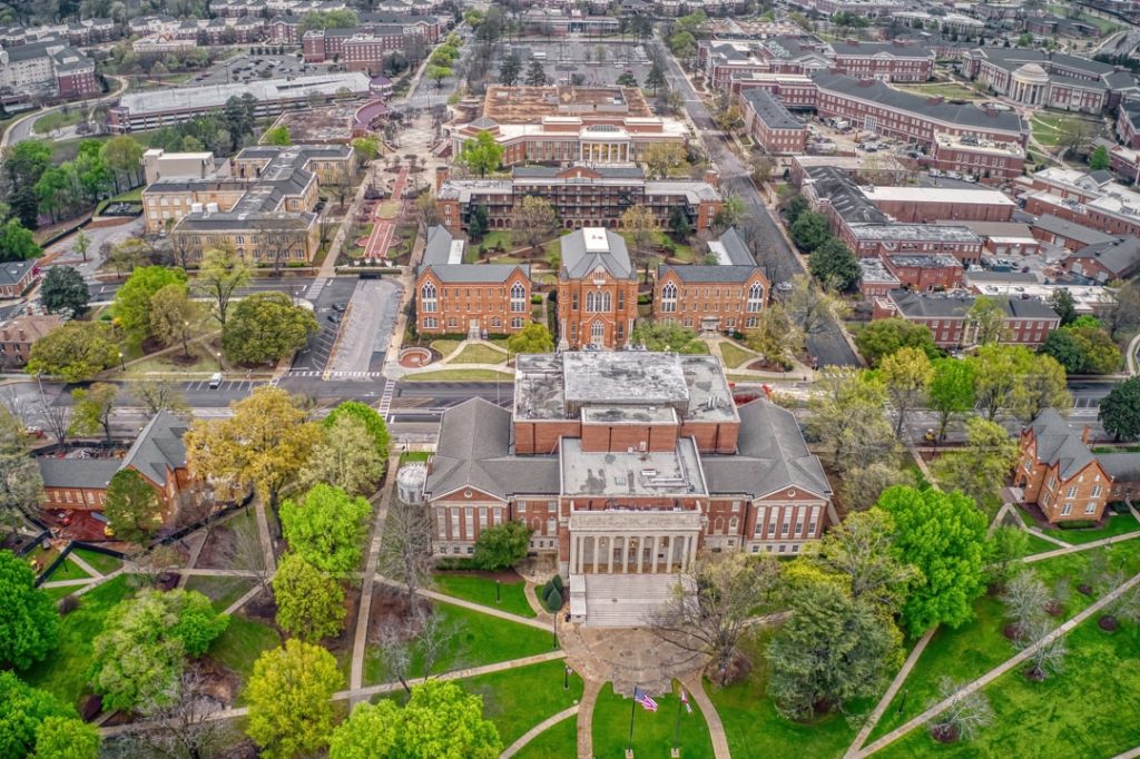 Luchtfoto van een bakstenen bibliotheekgebouw met zuilen omringd door groene campusgebouwen in Tuscaloosa
