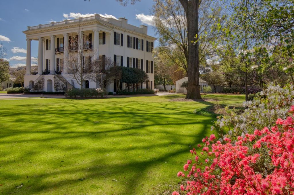 Wit zuilenhuis met veranda's op een groot gazon, met bloeiende roze azalea's op de voorgrond op de campus in Tuscaloosa