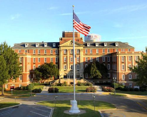 Bakstenen universiteitsgebouw met een grote Amerikaanse vlag op een vlaggenmast en een spandoek met de tekst Roll Tide