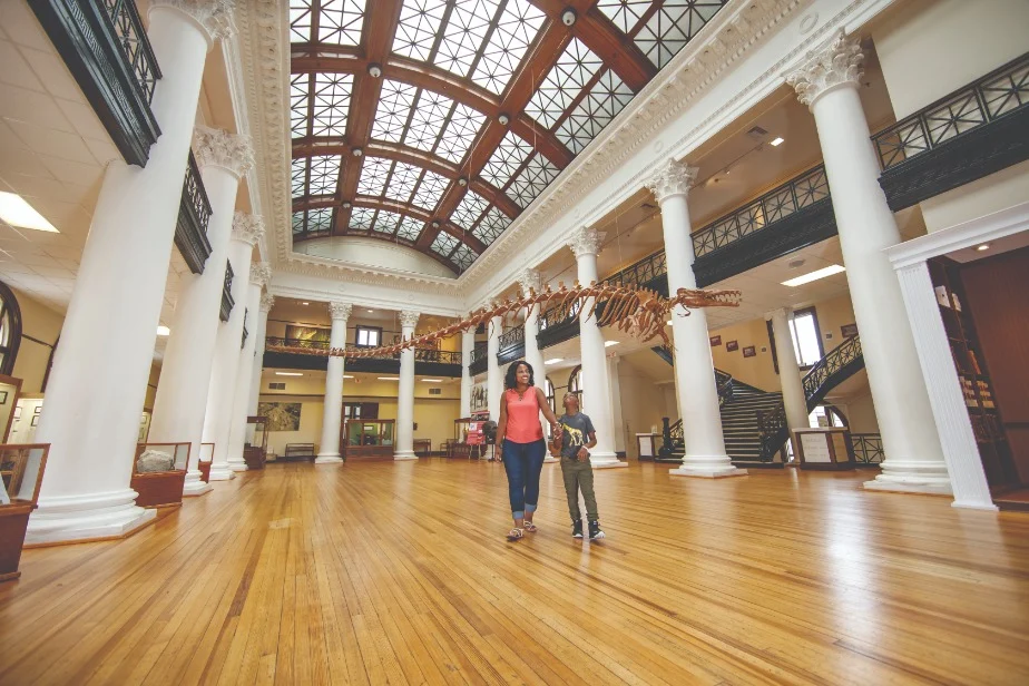 Een vrouw en een jongen lopen hand in hand door de grote hal van een natuurhistorisch museum in Tuscaloosa, met een opgehangen dinosaurusskelet en witte zuilen