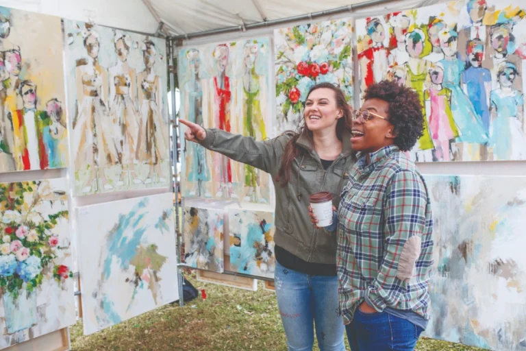 Twee lachende vrouwen bekijken kleurrijke schilderijen op een kunststand tijdens een buitenkunstfestival in Tuscaloosa