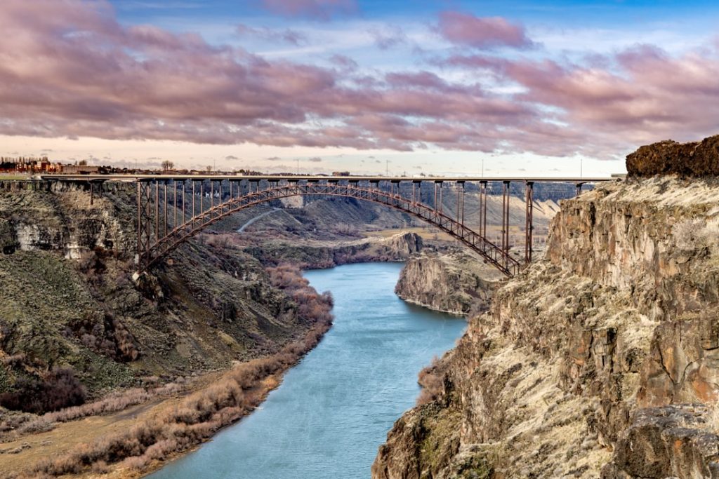 Stalen boogbrug overspant een diepe kloof met een blauwgroene rivier bij zonsondergang met roze wolken boven Twin Falls in Idaho