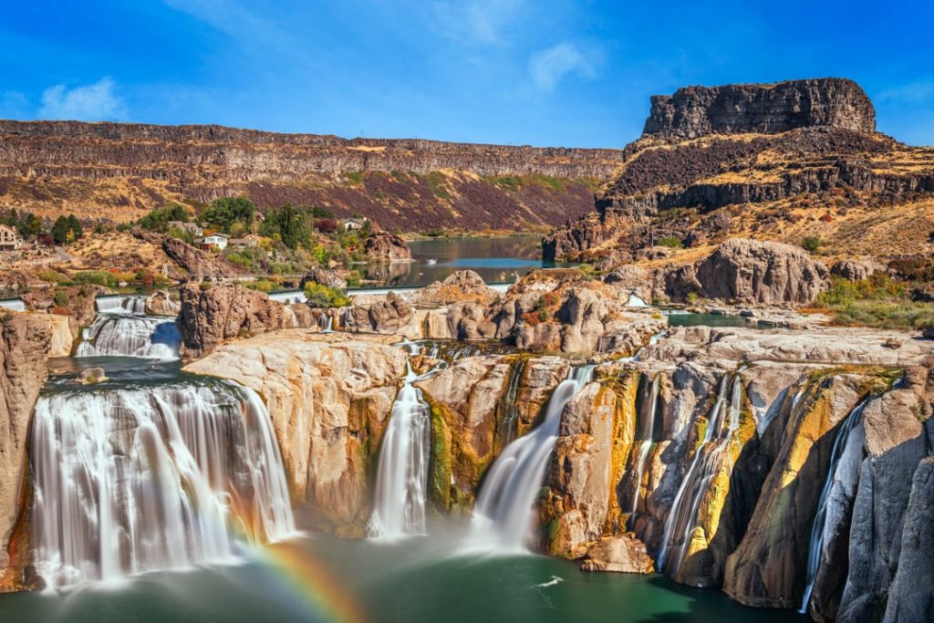 Brede waterval met meerdere stromen valt over gelaagde rotsen terwijl een regenboog zichtbaar is in de opspattende nevel