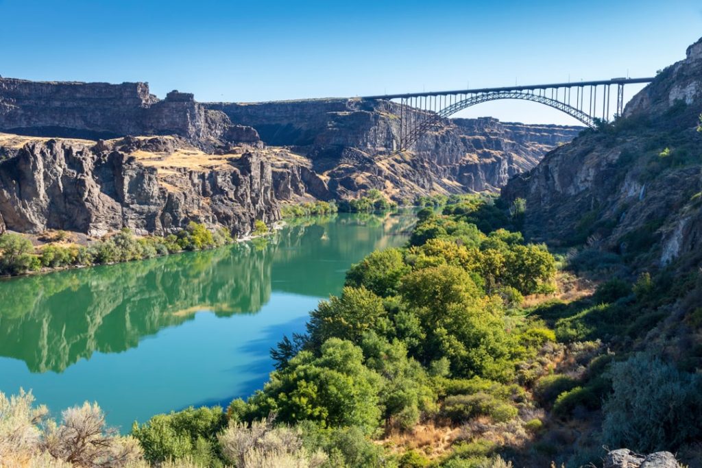 Groene rivier stroomt door een rotsachtige kloof met bomen langs de oever en een stalen boogbrug in de verte