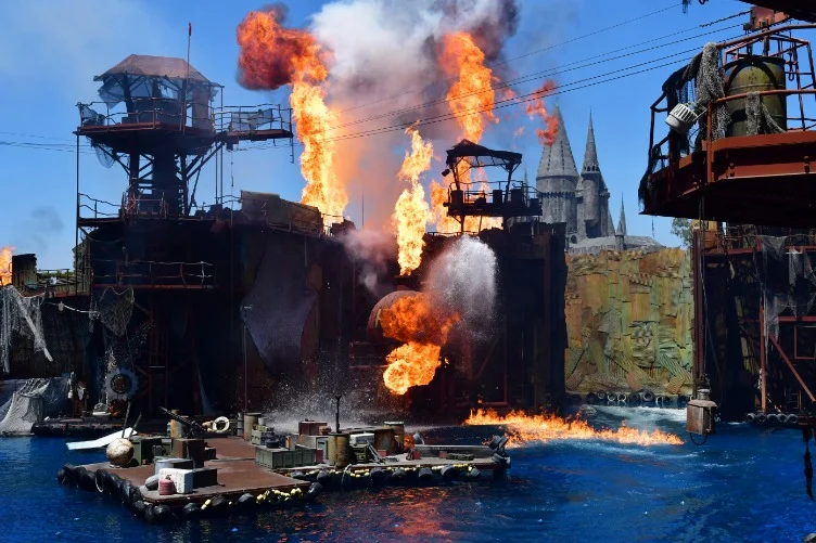 Grote vlammen en rook stijgen op boven een decor van vlotten en steigers tijdens de Waterworld stuntshow