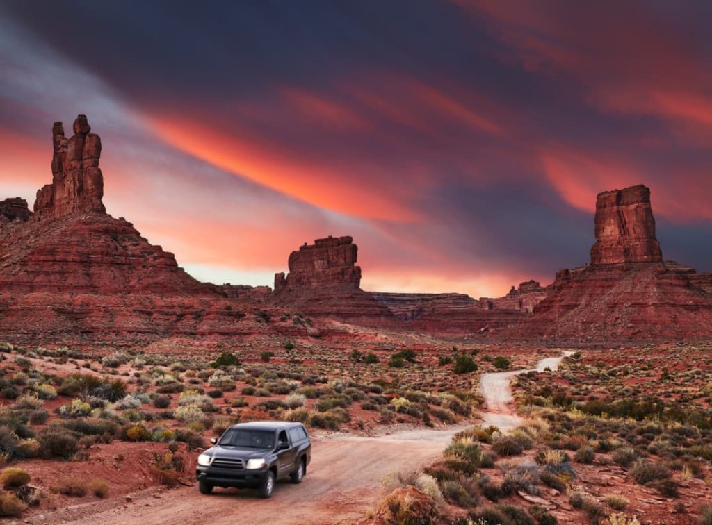 Een terreinwagen rijdt over een onverharde weg tussen indrukwekkende rode rotsformaties in Utah, met een gekleurde zonsonderganghemel op de achtergrond