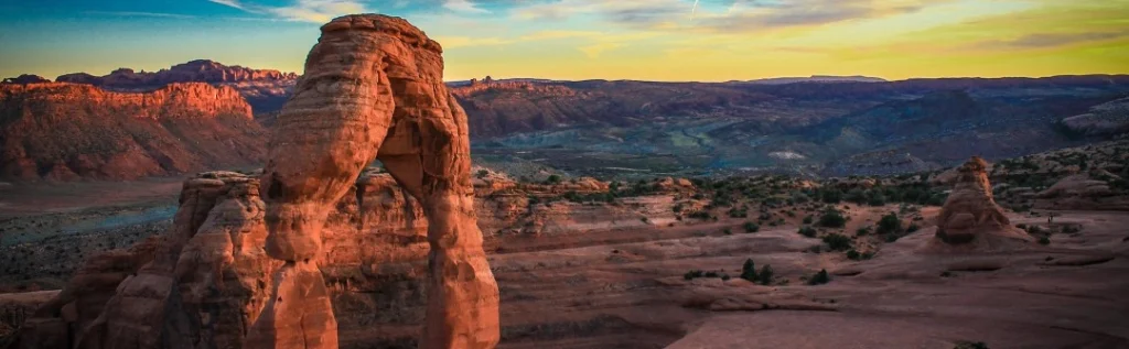 Vrijstaande rode zandstenen boog steekt af tegen een kleurrijke avondlucht met een uitgestrekt canyonlandschap op de achtergrond