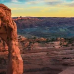 Vrijstaande rode zandstenen boog steekt af tegen een kleurrijke avondlucht met een uitgestrekt canyonlandschap op de achtergrond