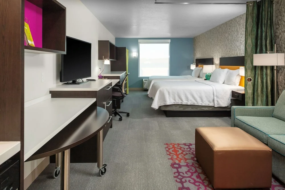 Ruime hotelkamer met een groot bed, televisie, bureau en een groene bank met kleurrijke kussens