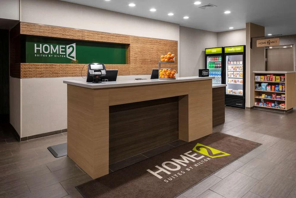 Houten receptiebalie van een hotel met een groen logobord, fruitmand en een kleine snackshop met koelkasten op de achtergrond