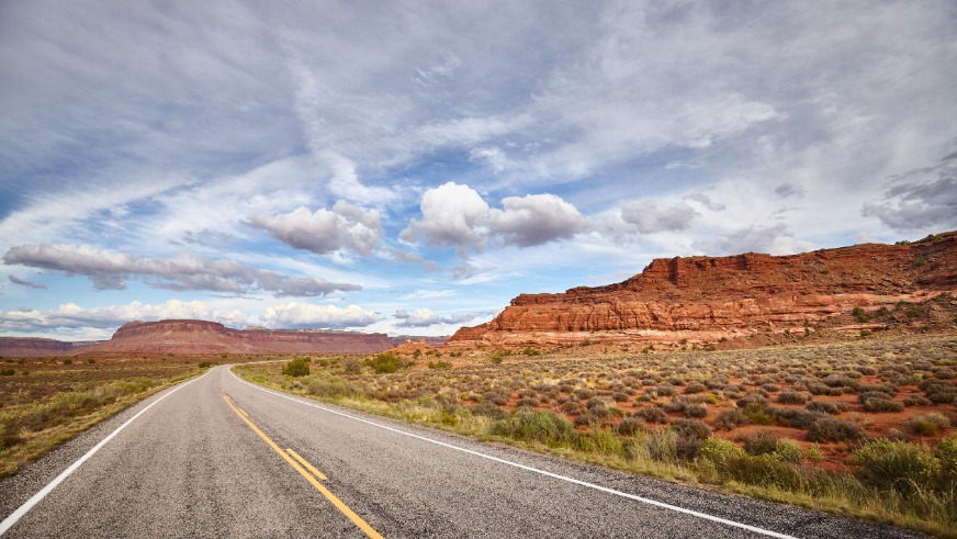 Een lege asfaltweg loopt door een dorre vlakte met struiken naar rode rotsformaties onder een bewolkte lucht in Utah