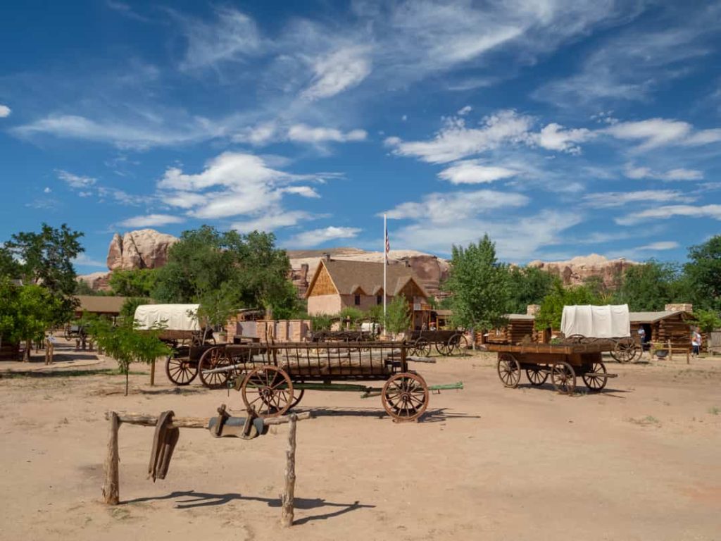 Verzameling houten huifkarren en wagenwielen op een zandvlakte bij een dorp in western stijl, met rode rotsen op de achtergrond in Utah