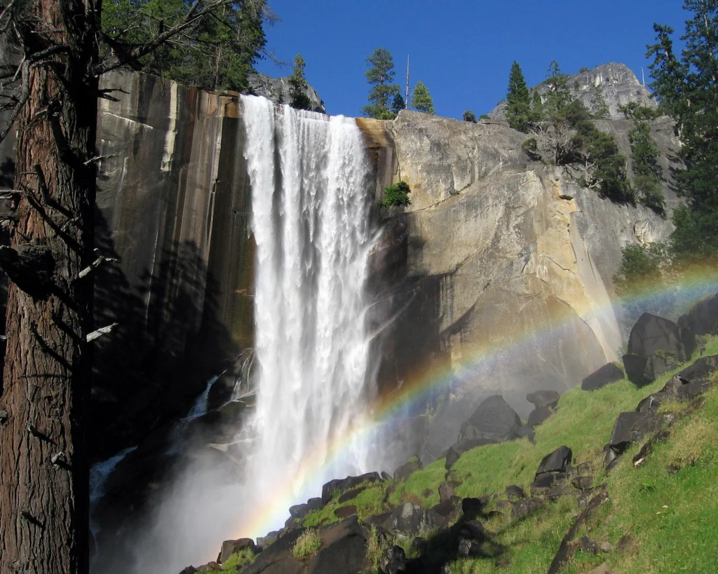 Krachtige waterval stort van een rotswand naar beneden met een dubbele regenboog erin, omringd door naaldbomen en rotsblokken