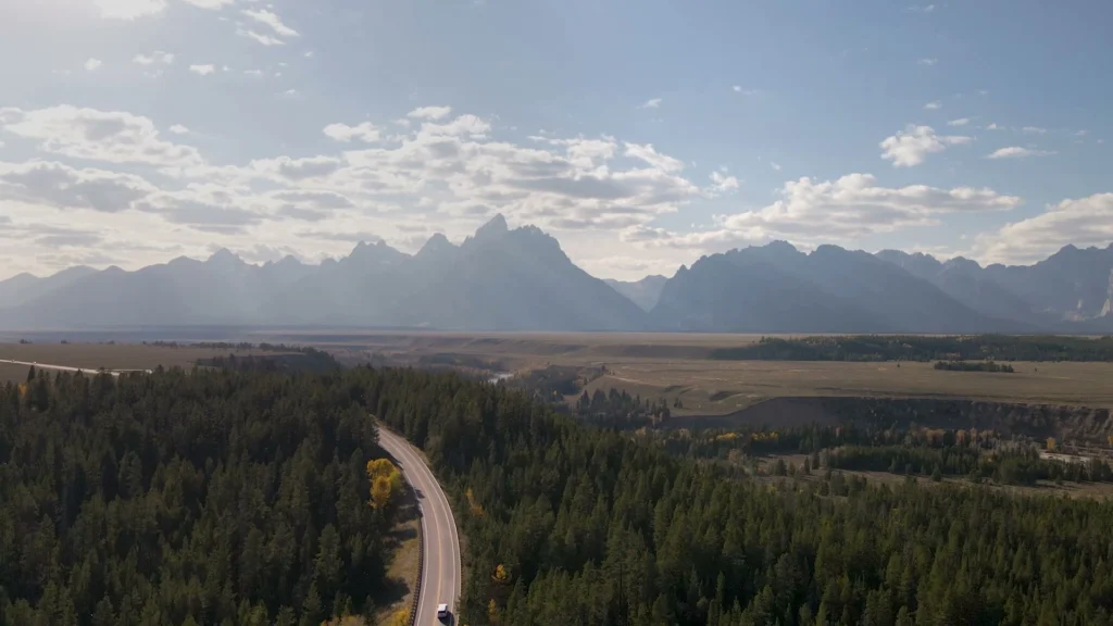 Een asfaltweg slingert door dennenbos met de bergketen van de Grand Tetons in de verte
