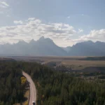 Een asfaltweg slingert door dennenbos met de bergketen van de Grand Tetons in de verte