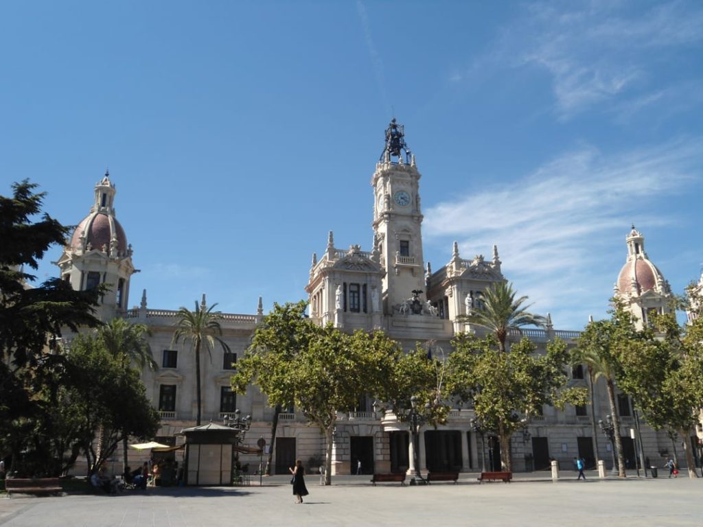 Monumentaal stadhuis van Valencia met koepels en een hoge klokkentoren met beeld aan een groot plein onder blauwe lucht
