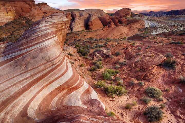 Golvende, roodwit gestreepte zandsteenrots op de voorgrond met woestijnstruiken en rotsen onder een roze avondlucht in Nevada