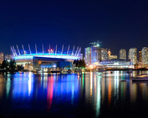 Wit stadion met blauw verlicht dak weerspiegeld in het water, omringd door verlichte wolkenkrabbers onder een donkerblauwe avondlucht