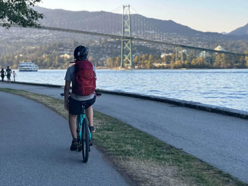 Fietser met rugzak rijdt over een pad langs het water in Stanley Park, met de Lions Gate Bridge zichtbaar
