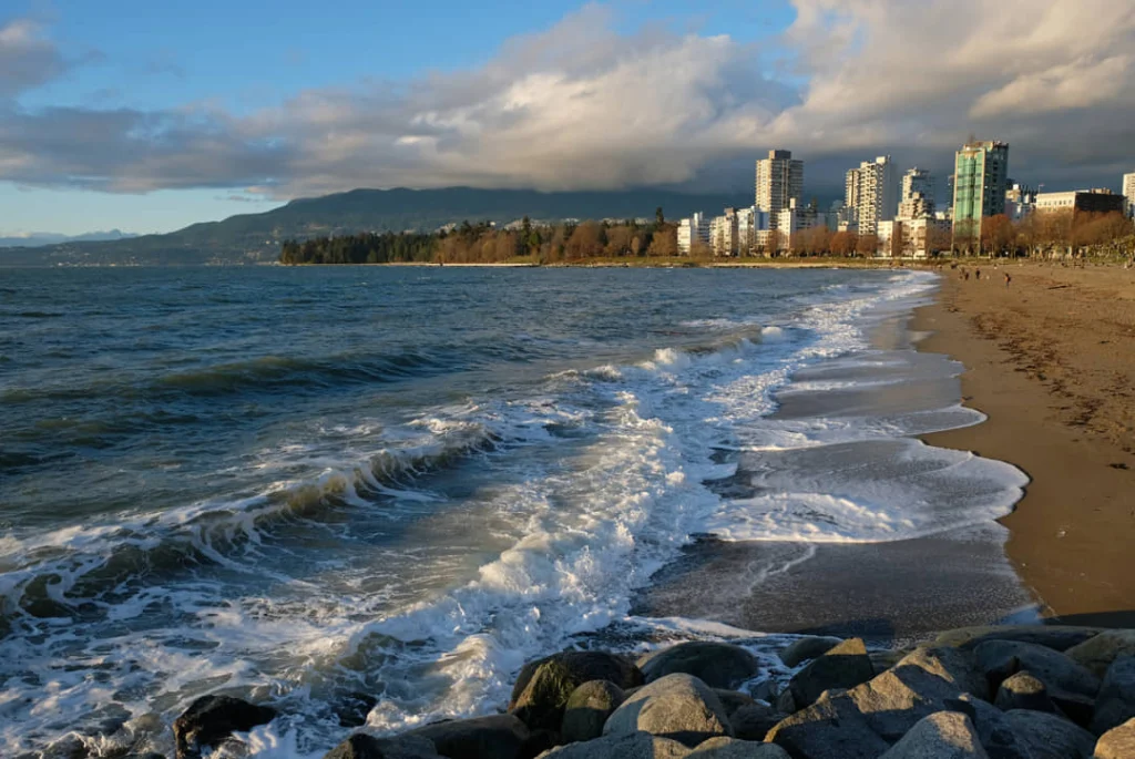 Schuimende golven spoelen over het zandstrand van Vancouver terwijl de skyline en bergen op de achtergrond zichtbaar zijn