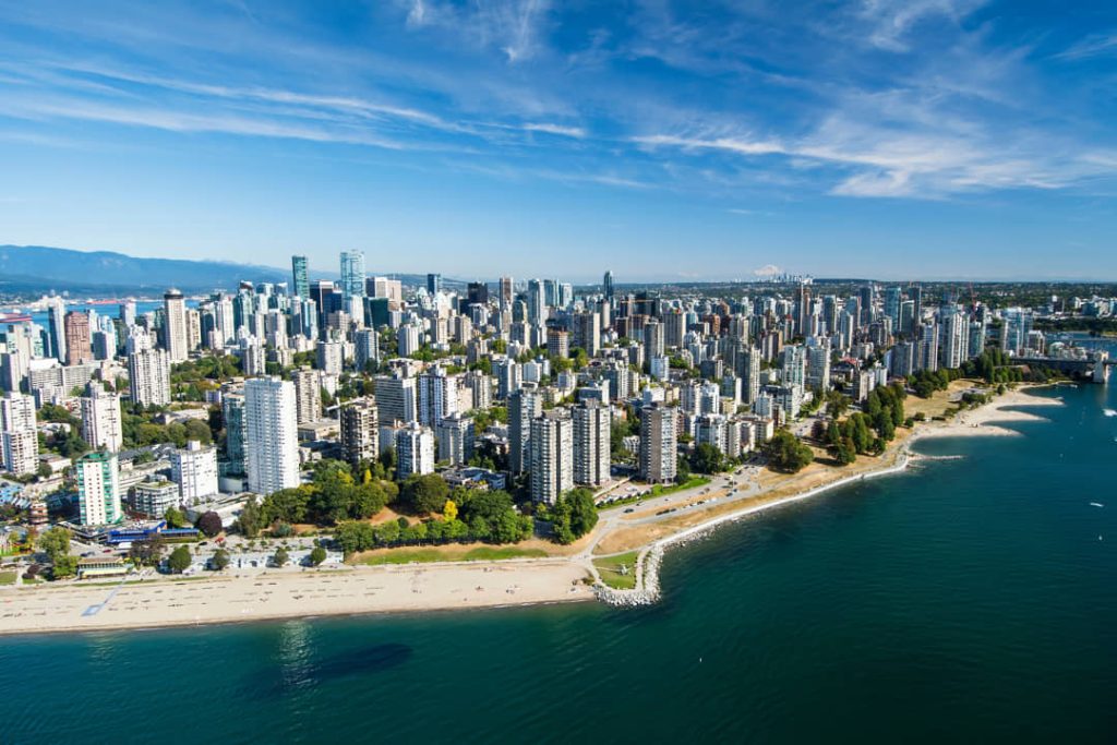 Luchtfoto van de kustlijn van Vancouver met een zandstrand, een park en talloze hoge appartementengebouwen langs het water onder een heldere lucht