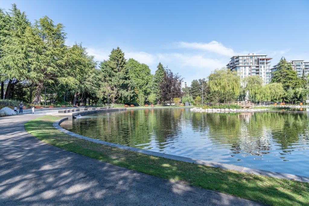 Rustige vijver in een stadspark met treurwilgen en wandelpaden, met een hoge woontoren op de achtergrond in Vancouver