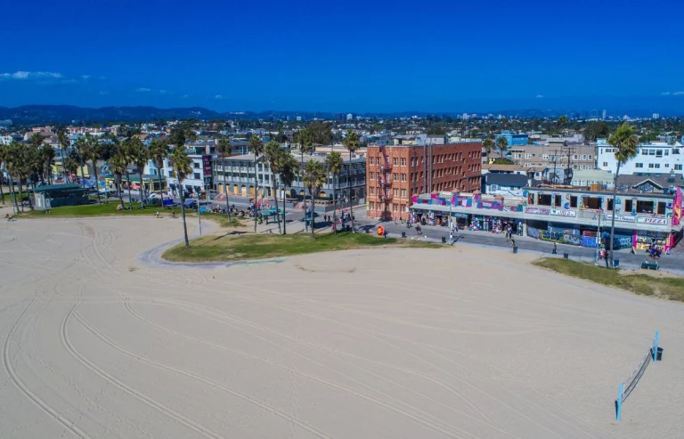 Luchtfoto van een breed zandstrand met daarachter gebouwen, palmbomen en de drukke boulevard van Venice Beach