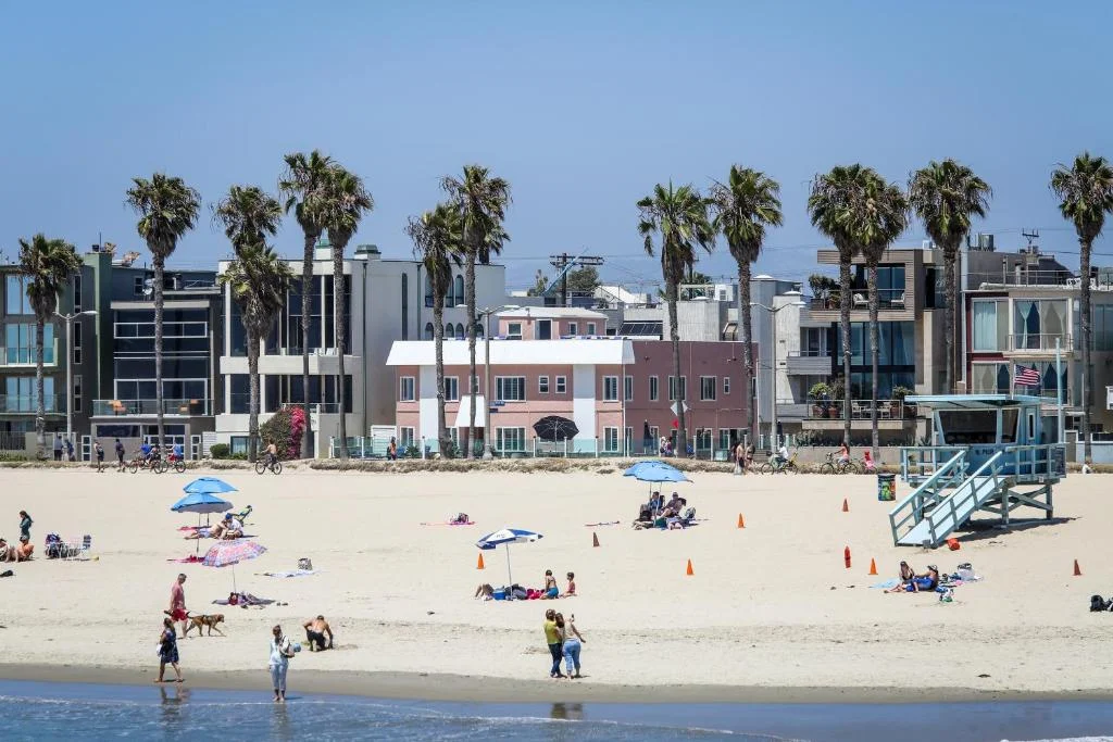 Strandgangers ontspannen op het zand met op de achtergrond hoge palmbomen, appartementen en een blauwe reddingstoren bij Venice Beach