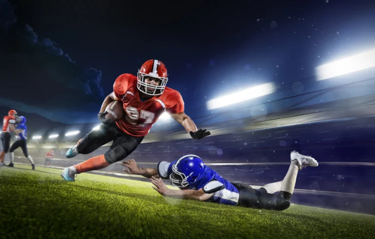 American football speler in rood tenue springt met de bal over een duikende tegenstander in blauw tenue onder stadionlicht