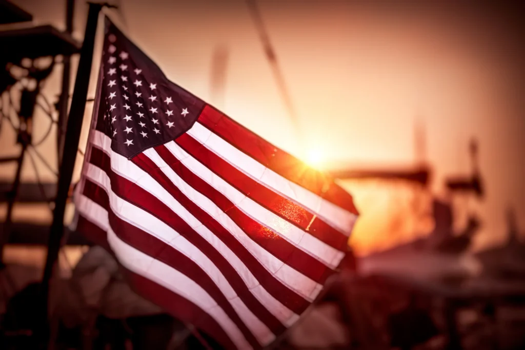 Amerikaanse vlag wappert op de voorgrond met silhouetten van installaties tegen een oranje avondlucht bij zonsondergang