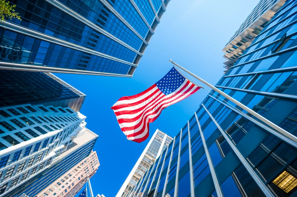 Amerikaanse vlag wappert aan een vlaggenmast tussen hoge glazen wolkenkrabbers gefotografeerd van onderaf tegen een blauwe lucht