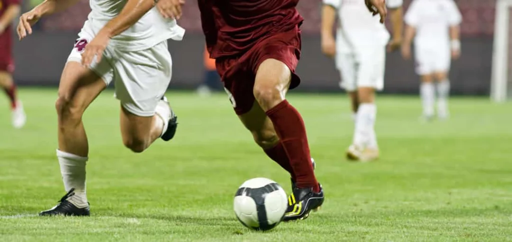 Close-up van de benen van twee voetballers in wit en bordeauxrood tenue tijdens een duel om de bal op het veld
