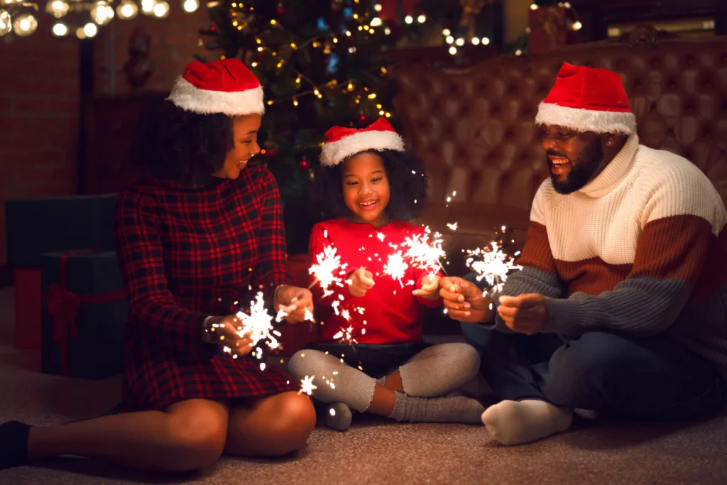 Ouders en dochter met rode kerstmutsen zitten met brandende sterretjes op de grond voor een verlichte kerstboom