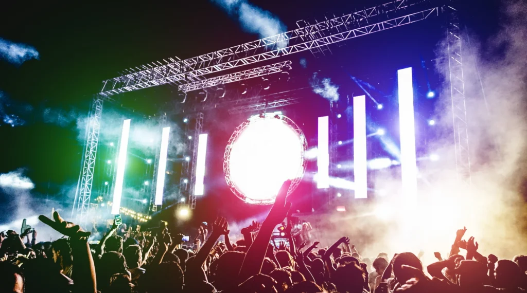 Juichende menigte met geheven armen voor een groot podium met groen roze en blauw licht en rook tijdens een muziekfestival