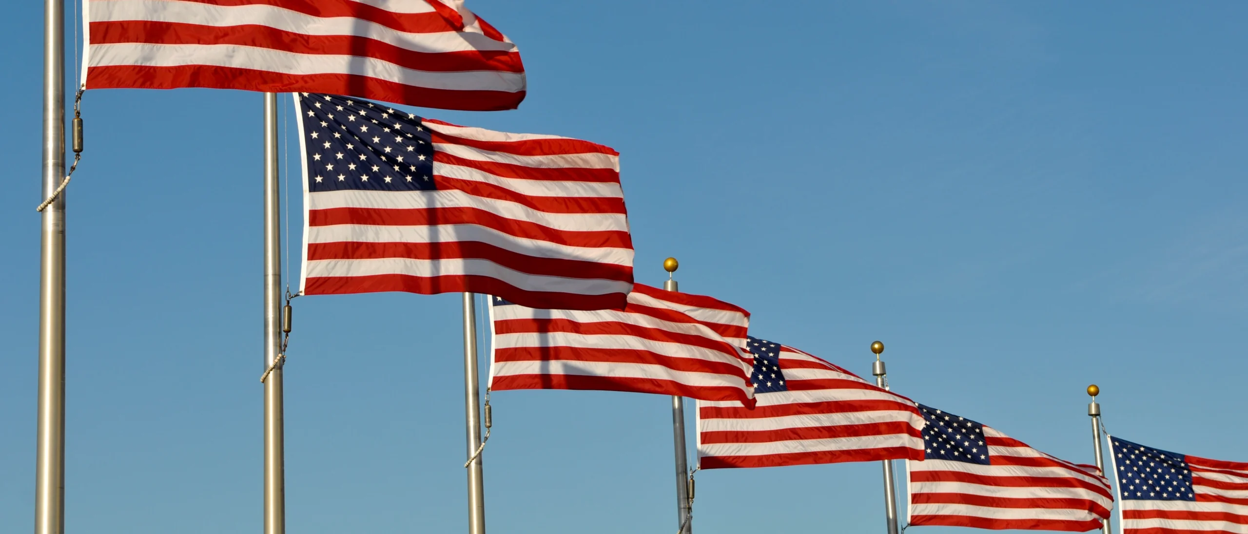 Meerdere Amerikaanse vlaggen aan vlaggenmasten wapperen op een rij tegen een strakblauwe lucht