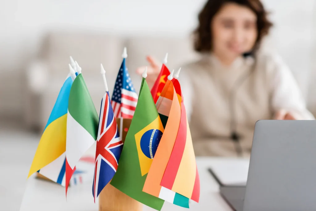 Kleine vlaggetjes van onder meer de Verenigde Staten Brazilië en het Verenigd Koninkrijk staan op een bureau naast een laptop met een vrouw op de achtergrond