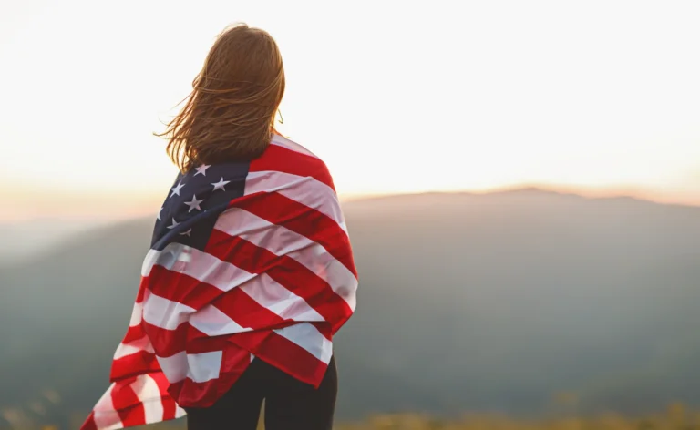 Vrouw met wapperend haar staat met een Amerikaanse vlag om haar schouders en kijkt uit over een berglandschap bij zonsondergang