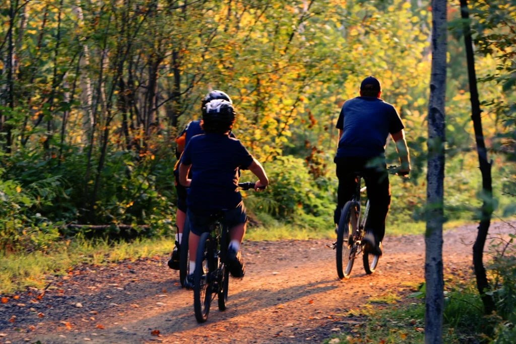 Ouder met twee kinderen fietsen achter elkaar over een onverhard bospad tussen bomen in geel en groen herfstlicht