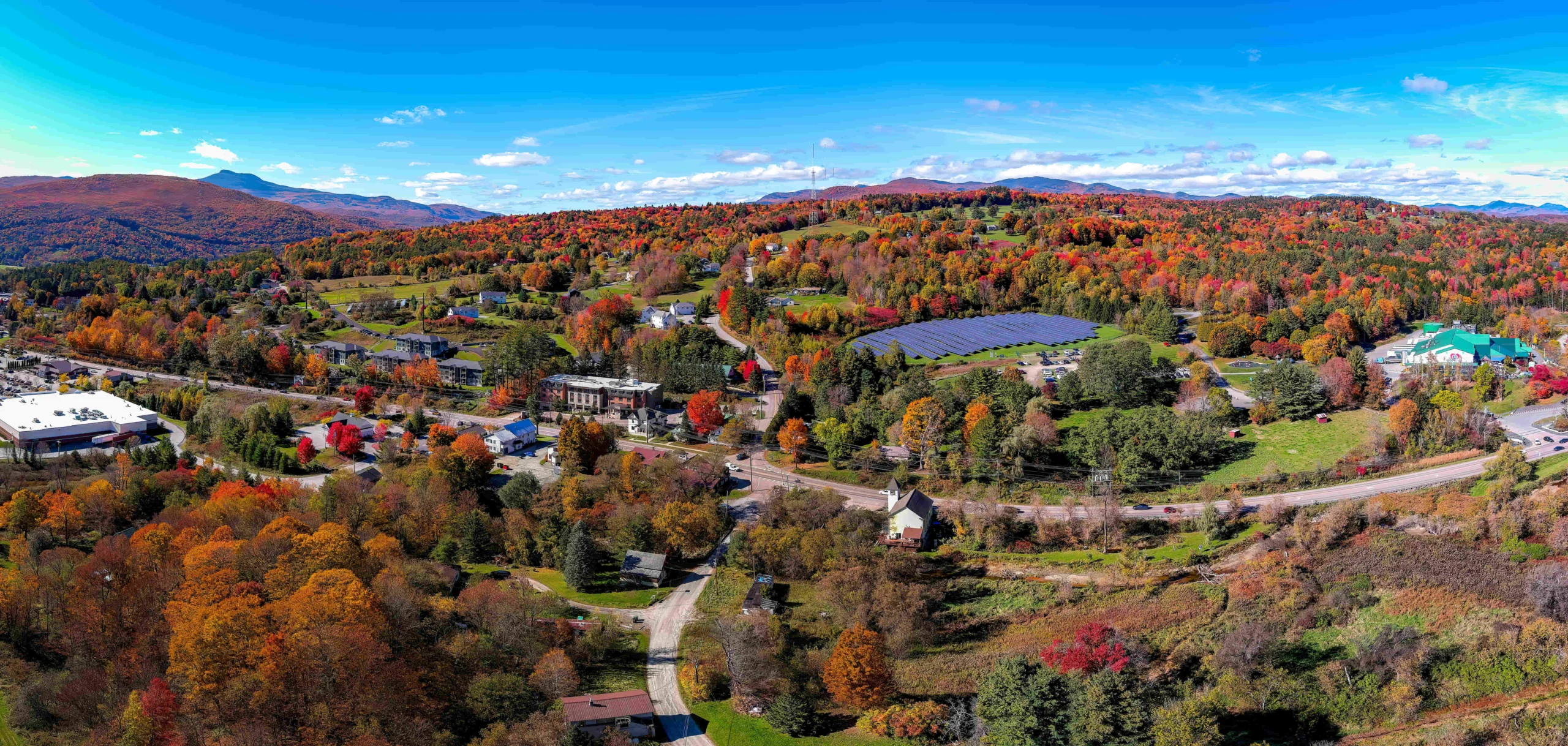 Luchtfoto van een dorp in Vermont omgeven door bossen in felle herfstkleuren en glooiende heuvels onder een blauwe lucht