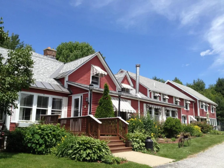 Lang rood houten gebouw met witte kozijnen, een overdekte veranda en een tuin met struiken in Vermont