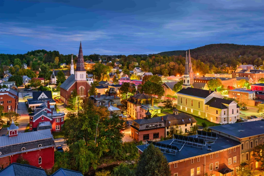 Luchtfoto van een verlicht stadje in Vermont met kerktorens en houten huizen tussen groene heuvels tijdens de schemering