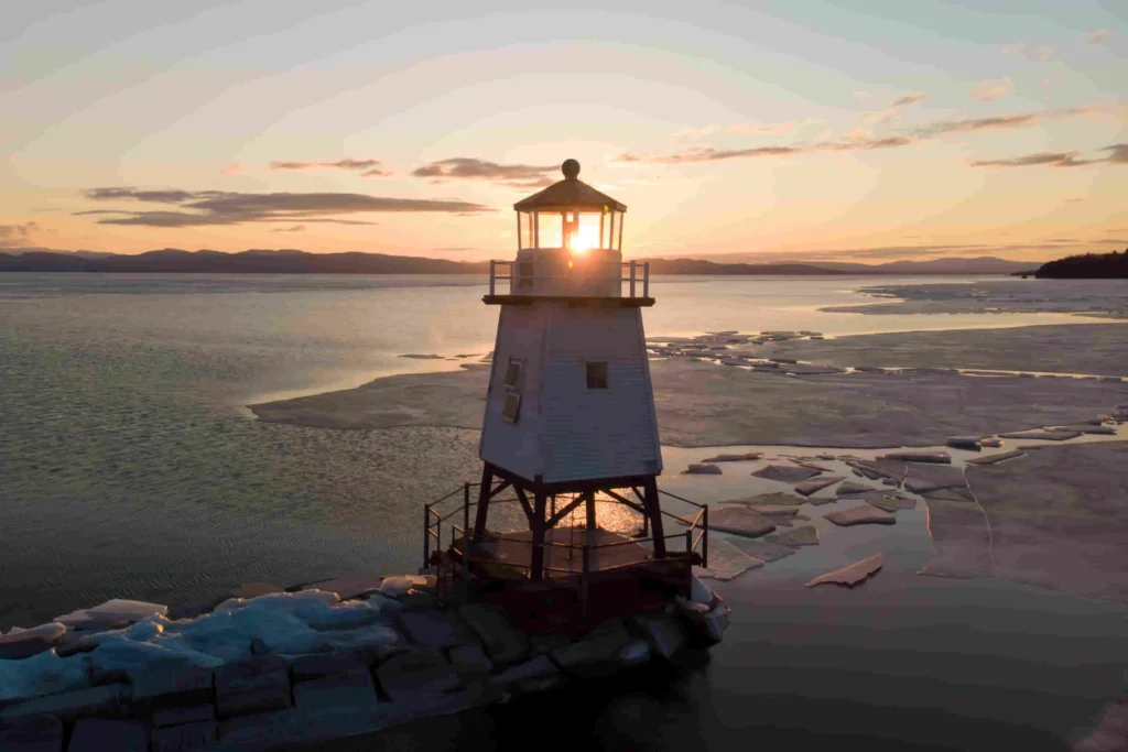 Een houten vuurtoren op een pier steekt uit boven een gedeeltelijk bevroren meer terwijl de zon door de lantaarnruimte schijnt, ergens in Vermont